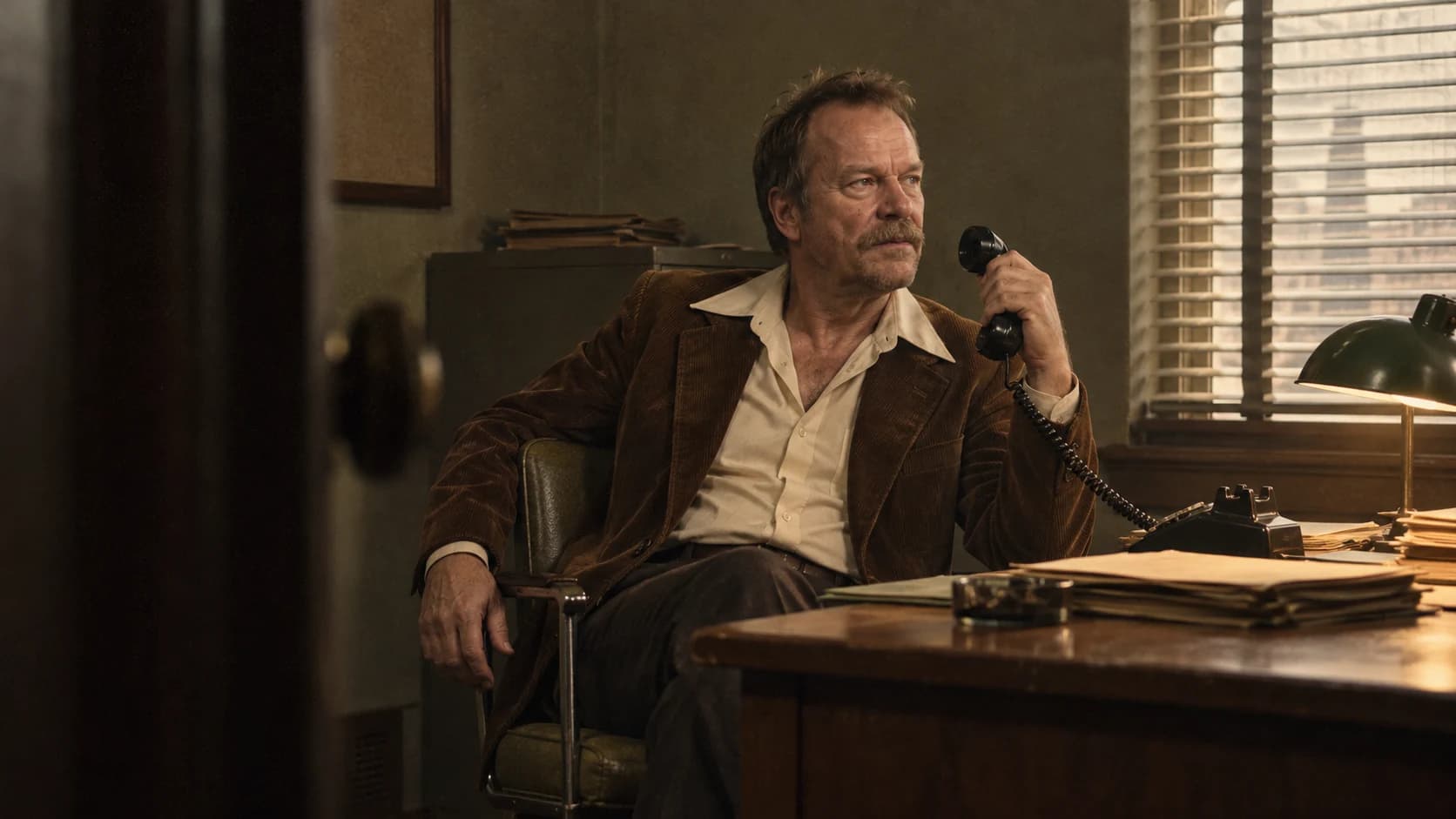 Brutalisti in a corduroy jacket holding a telephone in an olive-and-tobacco 1970s office.
