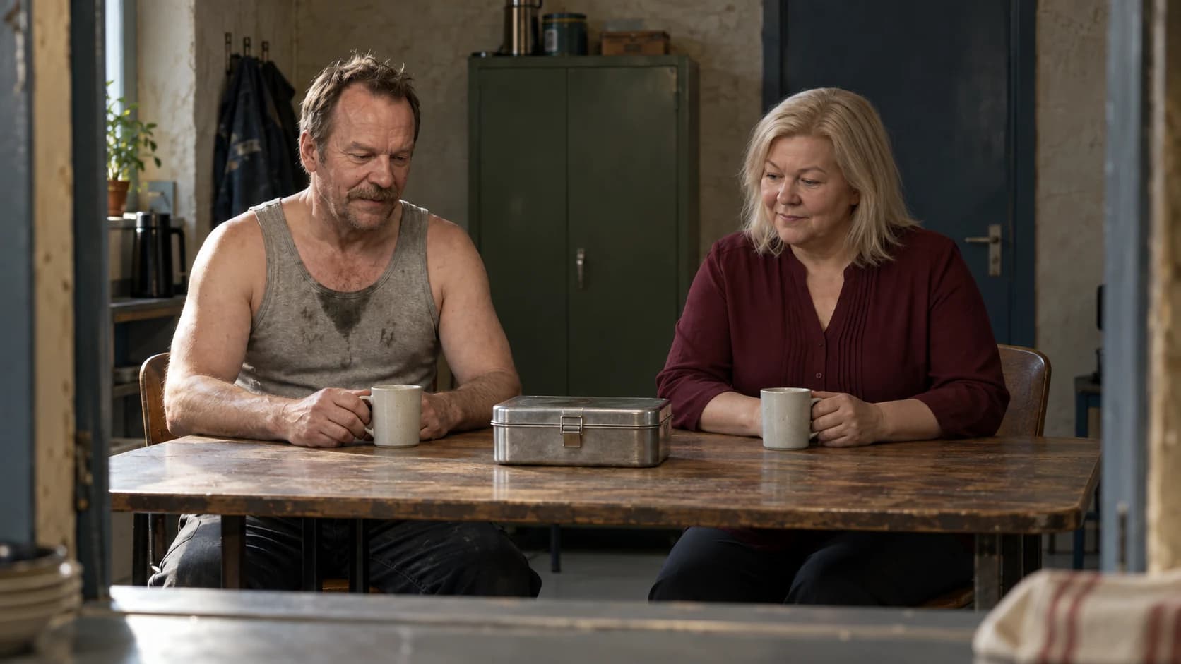 Brutalisti and Hilma sit at a depot break-room table with two cups and a metal lunchbox. Light on Brutalisti interpretation.