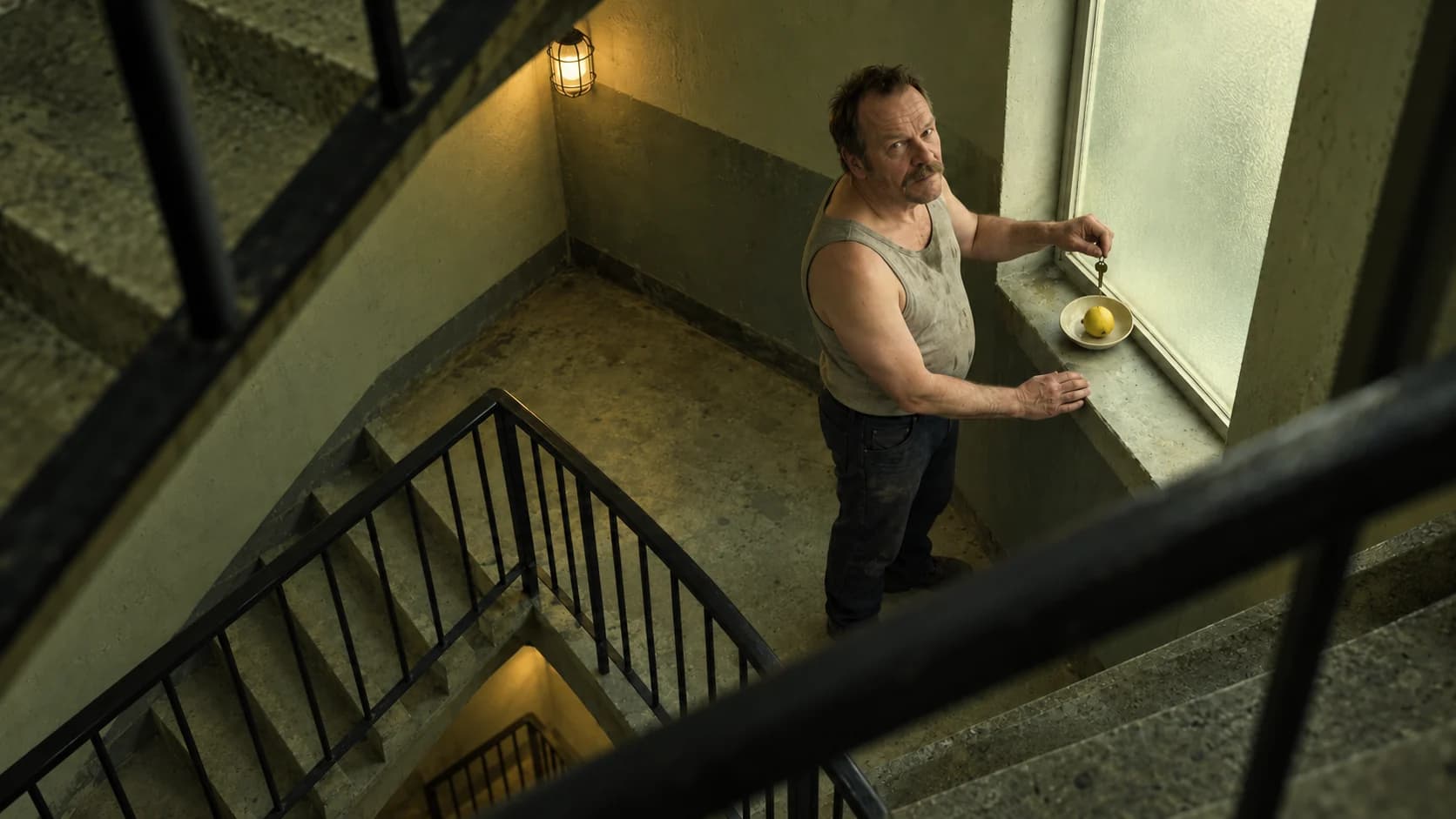 Seen steeply from above through diagonal concrete stairwell railings, Brutalisti holds a key suspended above a dish containing a lemon on the landing. Yellow-green interpretation.